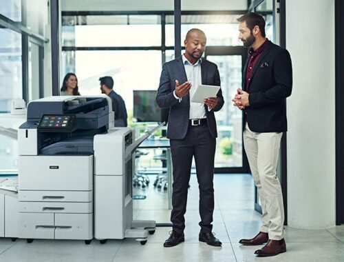 Two men in an office setting discussing something on a tablet beside a Xerox® AltaLink® C8200 Series Colour Multifunction Printer